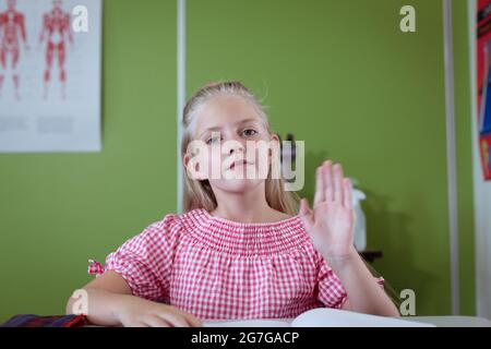 Porträt einer kaukasischen Schülerin, die im Klassenzimmer am Schreibtisch sitzt und die Hand hebt Stockfoto