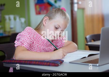 Kaukasische Schülerin am Schreibtisch im Klassenzimmer schreiben und mit Laptop Stockfoto