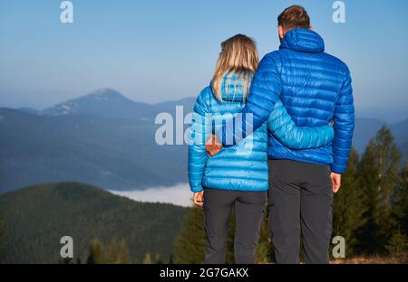 Rückansicht eines Wanderpaares, das sich um die Taille hält und vor sich bei Sonnenschein im Herbsturlaub eine fabelhafte Berglandschaft betrachtet. Stockfoto