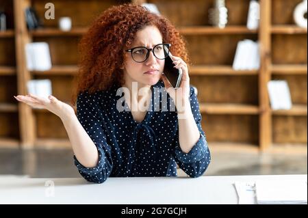 Verwirrt verwirrt verwirrt verwirrt Brünette Frau mit lockigen Frisur im Gespräch auf dem Handy und zum Ausdruck von Missverständnissen, macht eine hilflose Geste am Schreibtisch im Büro sitzen, Bücherregale im Hintergrund Stockfoto