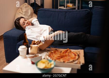 Kaukasische Frau schläft ein, nachdem sie Junk-Food im Wohnzimmer spät in der Nacht gegessen hat, während sie Essen zum Mitnehmen nach Hause geliefert hat. Leckere köstliche Mahlzeit Mittagessen Bestellung. Ungesunde Snacks Fastfood Stockfoto