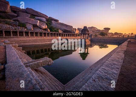 Blick auf Sonnenaufgang in Pushkarni, Sri Krishna Tank in Ruinen. südseite des Pools mit Schrein. Felsbrocken und Berge am Horizont. Hampi, Karnataka, Indien. Stockfoto