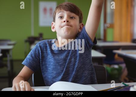 Aufgeregter kaukasischer Schuljunge im Klassenzimmer, der am Schreibtisch sitzt und die Hand hebt Stockfoto