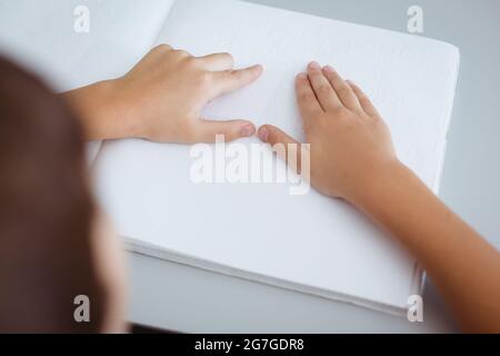 Blinder kaukasischer Schuljunge, der am Schreibtisch sitzt und mit den Fingern ein Braillebuch liest Stockfoto