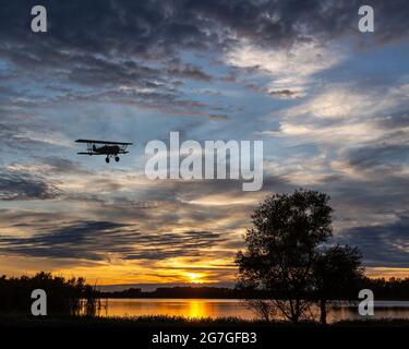 Ein Doppeldecker, der bei Sonnenuntergang tief über einem See fliegt Stockfoto