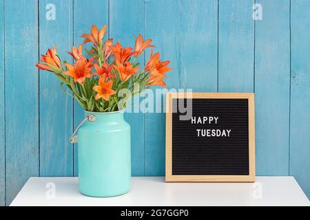 Happy Tuesday Worte auf schwarzem Briefbrett und Strauß oranger Blumen auf dem Tisch gegen blaue Holzwand. Konzept Hallo Dienstag. Stockfoto