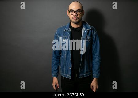 Porträt eines jungen gutaussehenden mexikanischen Glatzkopfes mit einer Brille, die vor einem grauen Hintergrund posiert Stockfoto