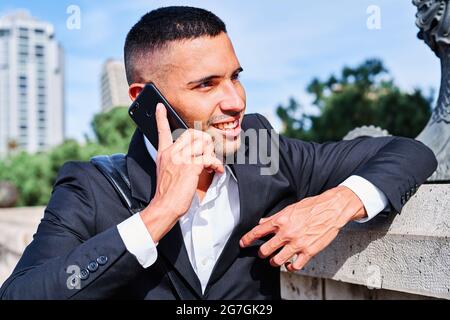 Selbstbewusster junger Mann in elegantem Anzug, der mit dem Handy spricht und lächelt, während er auf dem städtischen Platz in der Nähe der Skulptur steht Stockfoto