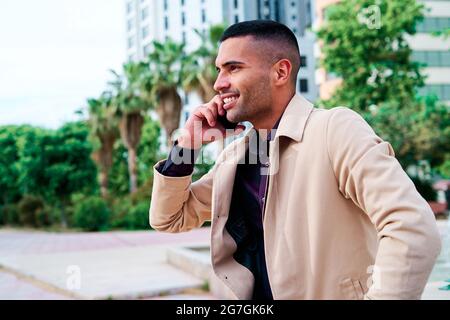 Positiver, gut gekleideter junger hispanischer Geschäftsmann, der auf dem Smartphone spricht und Nachrichten auf der städtischen Straße mit modernen Gebäuden im Hintergrund diskutiert Stockfoto