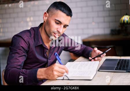 Seitenansicht eines gutaussehenden jungen hispanischen männlichen Unternehmers, der Informationen auf dem Smartphone überprüft und Notizen im Planer schreibt, während er am Tisch mit dem Schoß arbeitet Stockfoto