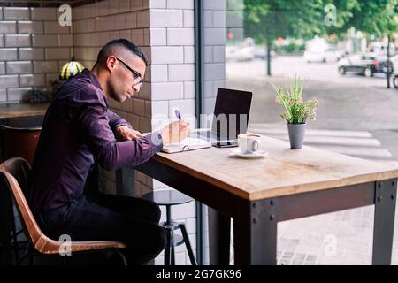 Serious hispanischen männlichen Manager in violettem Hemd und Brille tippen auf Netbook-Tastatur, während sie am Tisch hinter dem Fenster sitzen und an Projekt in Ca Stockfoto