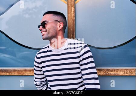 Positiver erwachsener Hispanic-Mann mit gestreiftem Pullover und stylischer Sonnenbrille, lächelnd und wegblickend, während er sich an einer dekorativen Glaswand auf der Stadtstraße lehnt Stockfoto