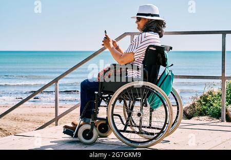 Seitenansicht einer behinderten Frau, die im Rollstuhl sitzt und auf dem Smartphone spricht, während sie den Sommerurlaub am Strand in der Nähe des Winkens verbringt Stockfoto