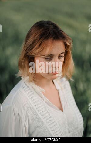 Ruhige junge Frau, die in altmodischer Bluse und Rock gekleidet ist und allein im hohen grünen Gras an bewölkten Sommertagen auf dem Land steht Stockfoto