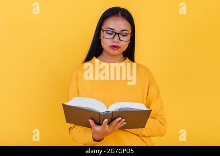 Clever asiatische Studentin in Brillen Lehrbuch lesen und Vorbereitung auf die Prüfung auf gelbem Hintergrund im Studio Stockfoto
