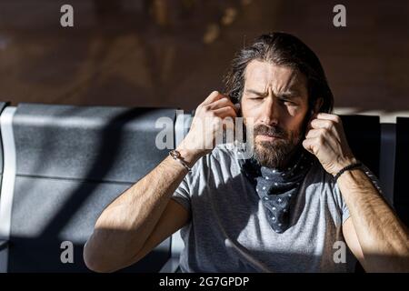 Der Mann im Hut am Flughafen im Wartezimmer, der auf seinen Flug wartet, setzt kabellose Kopfhörer an, um Musik zu hören Stockfoto