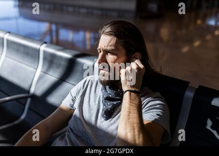 Der Mann im Hut am Flughafen im Wartezimmer, der auf seinen Flug wartet, setzt kabellose Kopfhörer an, um Musik zu hören Stockfoto