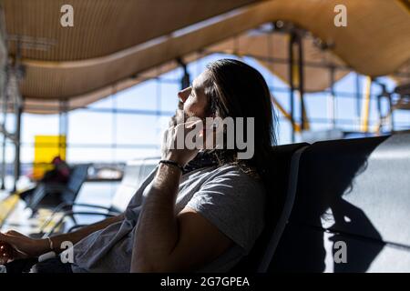 Der Mann im Hut am Flughafen im Wartezimmer, der auf seinen Flug wartet, setzt kabellose Kopfhörer an, um Musik zu hören Stockfoto