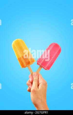 Rotes und gelbes Eis in Frauenhand auf blauem Hintergrund mit Kopierraum. Sommerhintergrund Stockfoto