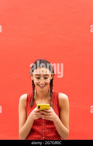 Fröhliche junge Frau in Zöpfen Frisur Browsen auf Smartphone auf rotem Hintergrund in der Straße stehen Stockfoto