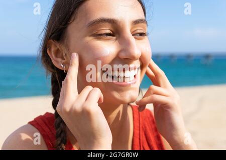 Lächelnde Frau, die am sonnigen Sommertag am Strand Sonnencreme auf das Gesicht aufgibt Stockfoto