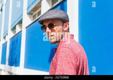 Junger bärtiger Kerl mit lässigem roten Polohemd und Mütze, der am Sommertag an der blauen Wand auf der Straße steht Stockfoto