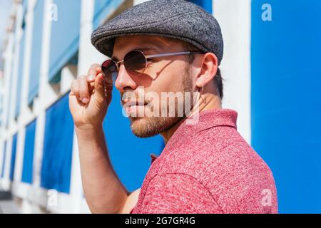 Junger bärtiger Kerl mit lässigem roten Polohemd und Mütze, der am Sommertag an der blauen Wand auf der Straße steht Stockfoto