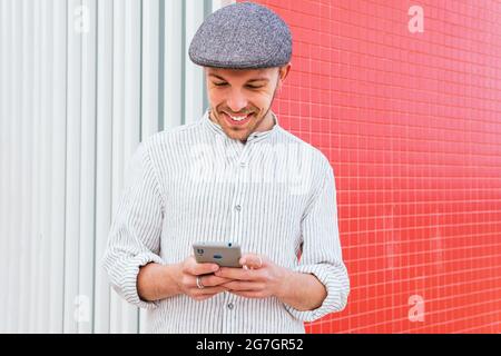 Inhalt junger bärtiger Hipster in legerem Poloshirt und Mütze, der im Sonnenlicht gegen die rote und weiße Wand steht und auf dem Handy surft Stockfoto