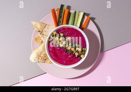Draufsicht auf appetitlichen Beetroot Hummus, garniert mit Kichererbsen, serviert auf zweifarbigem Hintergrund mit Brot und frischen Karotten- und Gurkenstöcken Stockfoto
