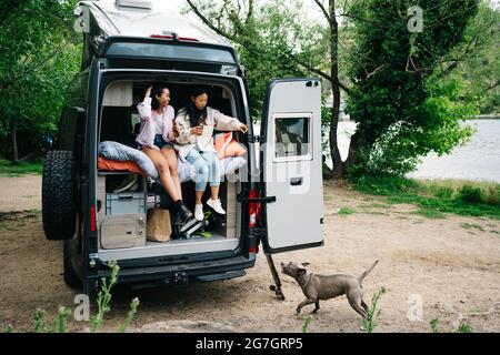 Reisende junge multirassische Freundinnen trinken Bier und geben Befehl an Hund, während Spaß haben und zusammen in Camper van in der Nähe des Flusses geparkt ausruhen Stockfoto