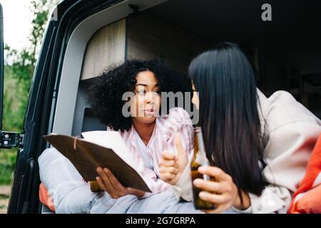 Fröhliche junge, multirassische Frauen trinken Bier und diskutieren Reiseroute mit Karte, während sie während der Sommerreise gemeinsam im Wohnmobil chillen Stockfoto