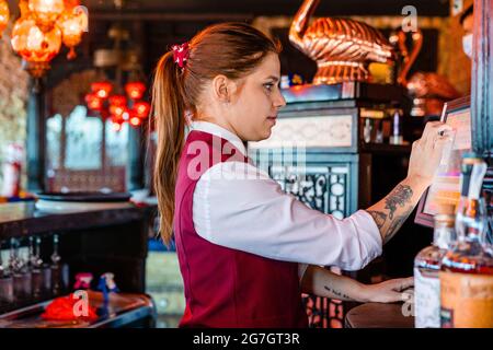 Seitenansicht der Barkeeperin, die die Kasse benutzt und den Bildschirm des Displays berührt, während sie in der Bar arbeitet Stockfoto