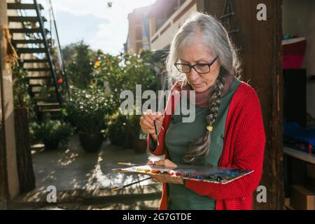 Fokussierte ältere Künstlerin mit Farbpalette und Pinsel, die an sonnigen Sommertagen im Hinterhof stehen Stockfoto