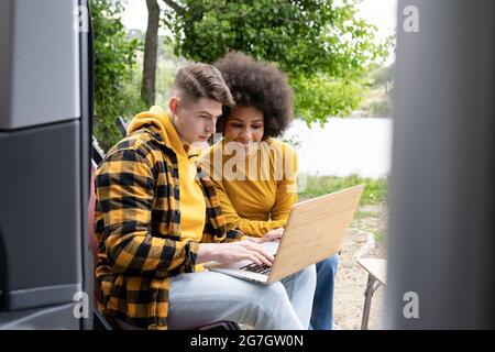 Junge, vielseitige Männer und Frauen in legerer Kleidung, die lächeln und Netbook durchsuchen, während sie während eines Roadtrip in die Natur in der Nähe des Van sitzen Stockfoto