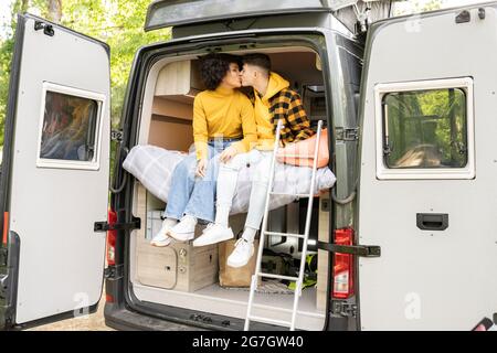 Verschiedene Männer und Frauen küssen sich gegenseitig, während sie auf dem Bett in einem modernen Van auf der Straße sitzen Stockfoto
