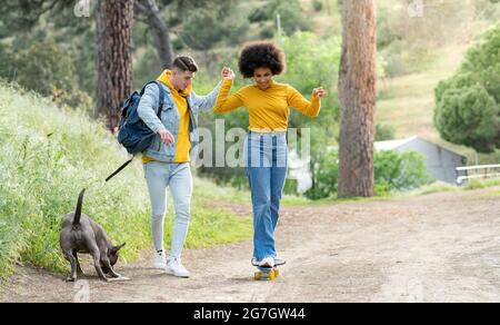 Ganzkörper junger Mann hält Hand und lehrt fröhlich schwarze Freundin Skateboard auf Landstraße in der Nähe von Hund in der Natur fahren Stockfoto