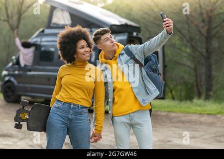 Verschiedene junge Männer und Frauen mit Skateboard, die Selfie in der Nähe von Van während der Road Trip am Sommertag in der Natur machen Stockfoto
