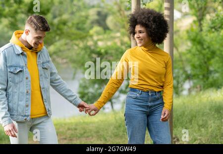 Entzückter, multiethnischer junger Mann und Frau, die sich die Hände halten, die an der Flussküste in der Natur spazieren gehen Stockfoto