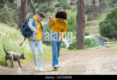 Ganzkörper junger Mann hält Hand und lehrt fröhlich schwarze Freundin Skateboard auf Landstraße in der Nähe von Hund in der Natur fahren Stockfoto