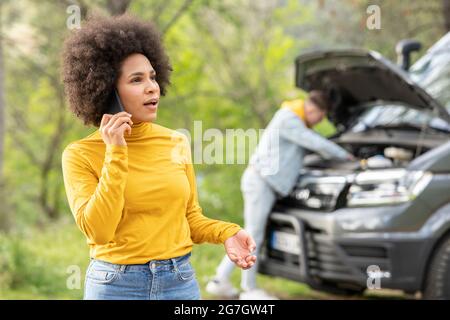 Schockierte schwarze Frau, die den Notdienst anruft, während ein junger Mann versucht, den Motor des Transporters während einer Autoreise auf dem Land zu reparieren Stockfoto