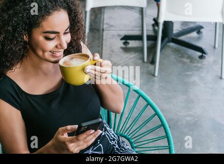 Von oben eine glückliche junge hispanische Frau, die auf dem Handy surft, während sie am Tisch des Straßencafés köstlichen Cappuccino genießt Stockfoto