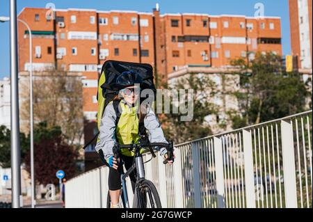 Fröhliche Kurierin mit Thermotasche, die Fahrrad auf der Brücke fährt, während sie Essen in der Stadt liefert Stockfoto