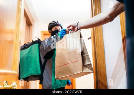 Delivery Frau mit thermischen Rucksack und Gesichtsmaske geben Papiertüte mit Lebensmitteln gesichtslosen Kunden durch Wohnungstür zu ernten Stockfoto