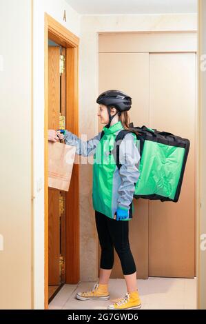 Seitenansicht der frohen Lieferfrau mit Thermorucksack, die dem Kunden durch die Wohnungstür eine Papiertüte mit Essen gab Stockfoto