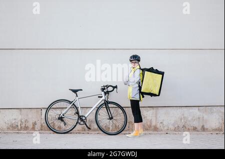 Ganzkörper-Kurierin mit Thermobeutel, die die Arme kreuzt und lächelnd auf die Kamera blickt, während sie vor dem modernen Gebäude neben dem Fahrrad steht Stockfoto
