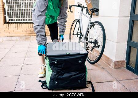 Anonyme Kurierin in uniform öffnende isolierte thermische Tasche auf gefliestem Boden in der Nähe der Tür während der Lieferung Stockfoto
