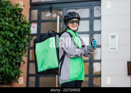Glückliche Geburt Frau mit Thermobeutel Blick auf die Kamera mit einem Lächeln und gestikulierenden Daumen nach oben vor dem Wohnhaus Stockfoto