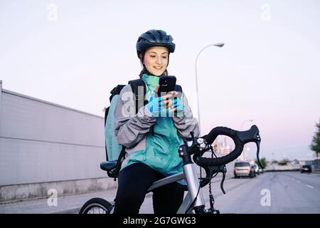 Junge, fröhliche Frau mit Thermotasche, die auf dem Smartphone mit GPS-Karte unterwegs ist, während sie auf dem Fahrrad sitzt, um Essen auf der Straße der Stadt zu liefern Stockfoto
