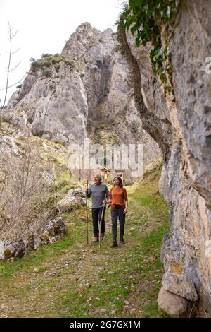 Ein ganzer Körper aktiver älterer Mann und Frau mit Holzstöcken, die auf einem steinigen Pfad zwischen blattlosen Bäumen und felsigen Bergen zusammenlaufen, während sie P erkunden Stockfoto