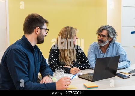 Begeisterte Mitarbeiter, die während eines Meetings am Arbeitsplatz am Tisch sitzen und über das Projekt sprechen, während sie sich gegenseitig anguckt Stockfoto